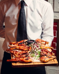 Waiter holding a wooden plate with grilled bbq chicken with onions greens sauce
