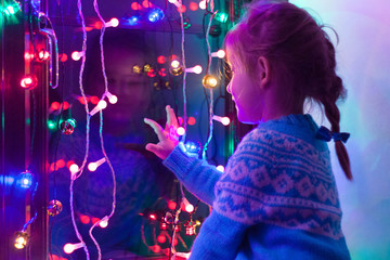 Happy little cute, beautiful girl 5 years old is sitting on the window and playing with Christmas lights and  Christmas decoration. Dark-blue theme.  - obrazy, fototapety, plakaty