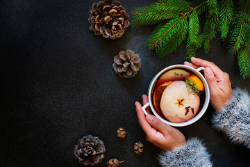 Female hands holding a mug of mulled wine. A warm drink made from red wine, citruses, fruits and spices. Winter, New Year, Christmas decorations. Top view, flat lay. Dark background. Copyspace