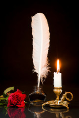 Red rose, feather in the inkwell and candle in the candlestick on a black background