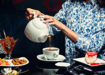 Woman pours in tea from teapot to a white cup, cheesecake, jam