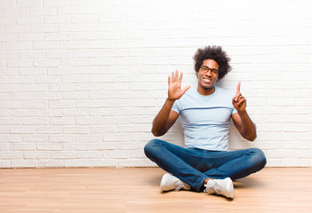 young black man smiling and looking friendly, showing number six or sixth with hand forward, counting down sitting on the floor at home