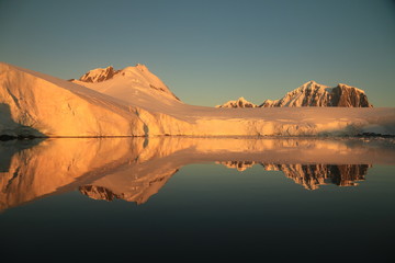 ośnieżone skalne wybrzeże antarktydy odbijające się w gładkiej tafli wody o zachodzie słońca © KOLA  STUDIO