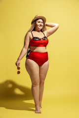 Young caucasian plus size female model's preparing for beach resort on yellow background. Woman in red swimsuit, hat and sunglasses. Concept of summertime, party, body positive, equality and chill.