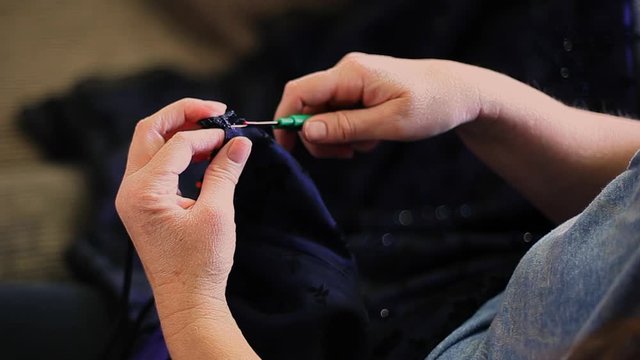 Dry, cracking, lizard like hands of a middle aged woman rip stitches from a formal dress in attempts to alter it to fit. Taking top beaded accessory off the bodice of a dress.