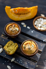 Pumpkin muffins on a dark wooden and half a broken cupcake and a slice of pumpkin in the background and seeds