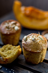 Pumpkin muffins on a dark wooden and half a broken cupcake and a slice of pumpkin in the background and seeds