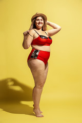 Young caucasian plus size female model's preparing for beach resort on yellow background. Woman in red swimsuit, hat and sunglasses. Concept of summertime, party, body positive, equality and chill.