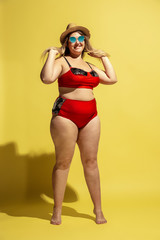 Young caucasian plus size female model's preparing for beach resort on yellow background. Woman in red swimsuit, hat and sunglasses. Concept of summertime, party, body positive, equality and chill.