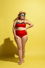 Young caucasian plus size female model's preparing for beach resort on yellow background. Woman in red swimsuit, hat and sunglasses. Concept of summertime, party, body positive, equality and chill.