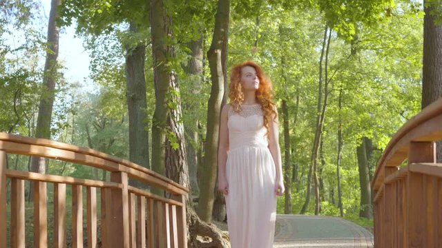 Young readhead girl walking in the park. Pretty caucasian woman in long light dress resting in the sunrays.