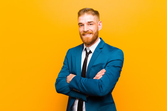 young red head businessman smiling to camera with crossed arms and a happy, confident, satisfied expression, lateral view against orange background