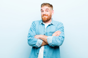 young red head man shrugging, feeling confused and uncertain, doubting with arms crossed and...