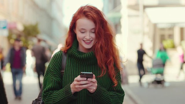 Charming young woman in green sweater walks in the city center smiles uses phone feel happy positive emotion outside