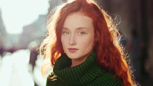 Portrait of seriously young red hair woman in green sweater looking at camera in the city center the sun is shining at the background