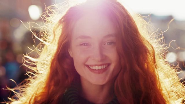 Portrait of beautiful young woman with curly red hair looking and smiling at camera sun is shining at the background positive emotion
