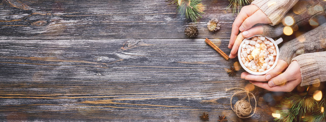 Woman's and man's hands holding the cup of hot cocoa with marshmallows on wooden background. Banner with winter hot drink.
