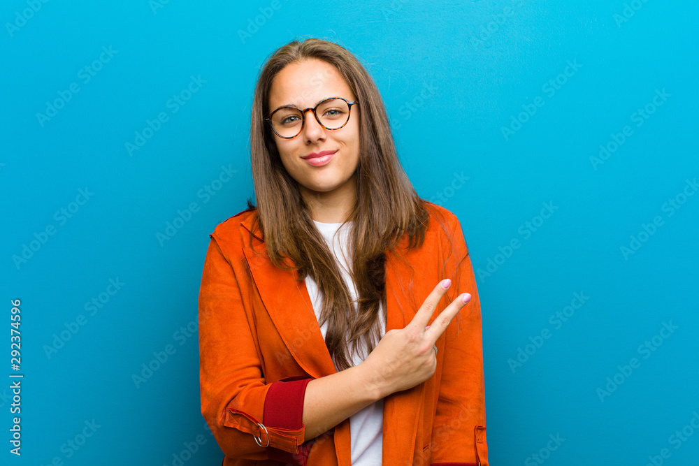 Wall mural young woman feeling happy, positive and successful, with hand making v shape over chest, showing vic