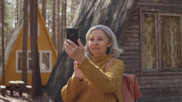 Medium shot of grey-haired elderly woman standing near cozy houses in forest and making photos