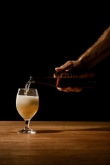 partial view of man pouring beer from bottle in glass on wooden table isolated on black