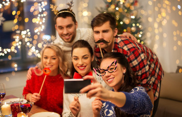 Obraz na płótnie Canvas holidays and celebration concept - happy friends with party props taking selfie by smartphone at home christmas dinner