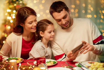 holidays, family and technology concept - happy mother, father and little daughter with smartphone having christmas dinner at home