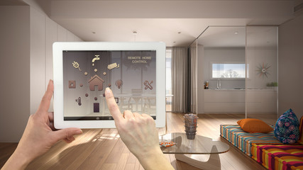 Smart remote home control system on a digital tablet. Device with app icons. Minimalist modern bright living room with kitchen in the background, architecture interior design