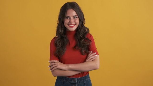 Pretty smiling brunette girl in red top happily looking in camera with hands crossed over yellow background