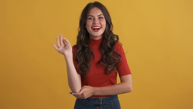 Cheerful brunette girl in red top joyfully waving hand hello on camera over yellow background. Greeting gesture
