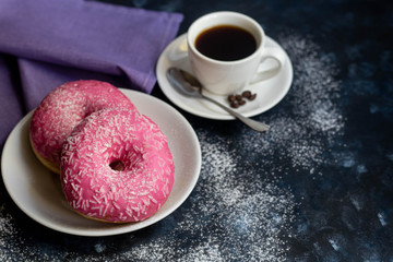 Fragrant, hot coffee. Next to a plate are donuts with a pink brush and white sprinkle. Everything is located on a dark background. 