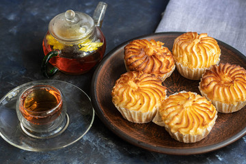 On a brown clay plate are cupcakes with cream filling decorated with raspberries. In the background is floral tea with ginger and lime. Everything is located on a dark background. 