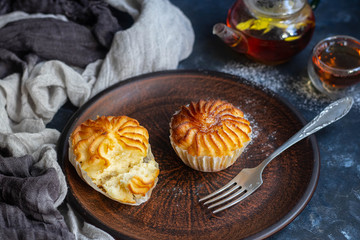 On a brown clay plate are cupcakes with cream filling decorated with raspberries. In the background is floral tea with ginger and lime. Everything is located on a dark background. 