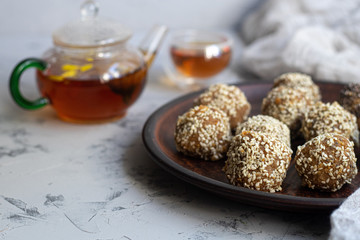On a brown clay plate are Indian sweets Lada. They are sprinkled with sesame seeds. In the background is flower tea with ginger and lime. Everything is located on a gray background decorated in granit