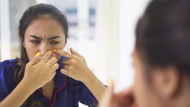 Beautiful teenage girl squeezing pimples on her face while looking at the mirror in the bedroom at home. Shot in 4k resolution