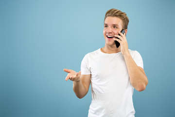 Caucasian young man's half-length portrait on blue studio background. Beautiful male model in shirt. Concept of human emotions, facial expression, sales, ad. Talking on phone, looks happy.