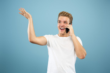 Fototapeta premium Caucasian young man's half-length portrait on blue studio background. Beautiful male model in shirt. Concept of human emotions, facial expression, sales, ad. Talking on phone, looks happy.
