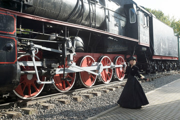 Steampunk girl in a black dress and hat near an old steam locomotive and large iron wheels. Blond beauty. Vintage portrait of the last century, retro journey.