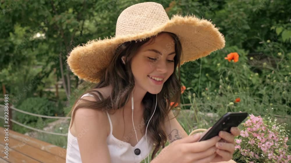 Poster pleased cute young brunette woman in straw hat listening music with earphones and chatting on smartp