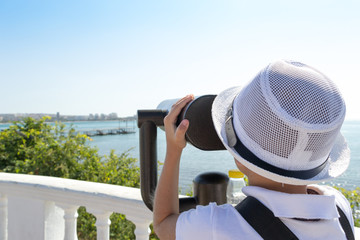 View from behind of little boy looking through a stationary binoculars at the beautiful seascape during his journey. Sunny day. Travel in vacation.