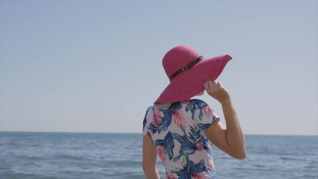 Lonely elegant woman in blue swimsuit and pink and hat standing by the sea and looking at the horizon