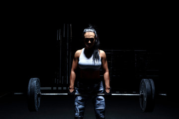 Woman bodybuilder make her workout with weights - barbell. Light from above, dark background, mysterious photo.