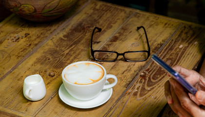 White cup of coffee with flower latte art and little milk mug with eyeglasses on wooden table background.