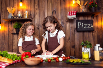 Happy little girls with rolling pin and spatula smiling and looking at camera while preparing delicious pizza in cozy kitchen together