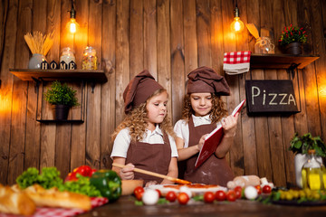Cute girls in aprons and hats reading recipe book together while cooking tasty pizza in cozy kitchen at home
