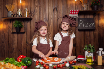 Happy little girls with rolling pin and spatula smiling and looking at camera while preparing delicious pizza in cozy kitchen together