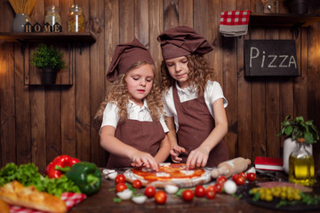 Happy little girls with rolling pin and spatula smiling and looking at camera while preparing delicious pizza in cozy kitchen together