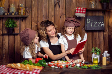 Smiling woman reading recipe book with girls in aprons and hats while cooking pizza in cozy kitchen at home together