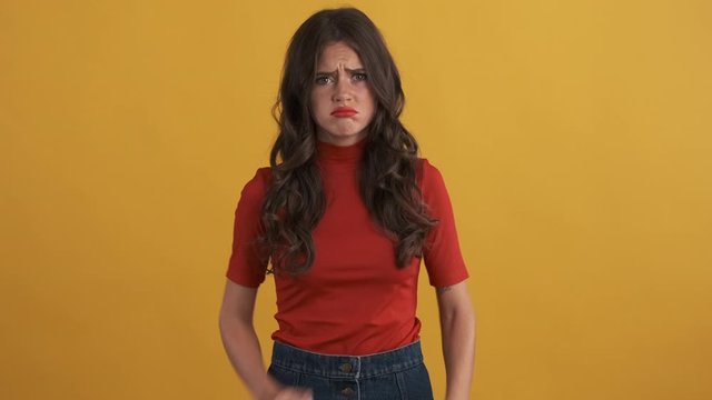 Upset moody brunette girl in red top offendedly posing on camera over yellow background