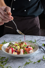 Chef serving sauce on the salad with melon, proshuto, arugula and italian cheese in a white plate. Spoon, fork on the grey marble background