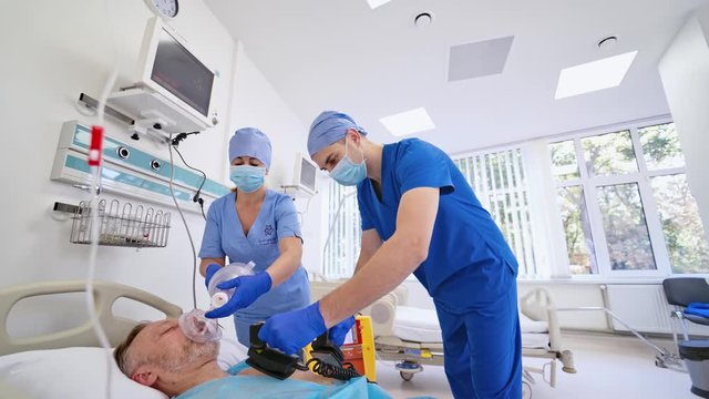 Male unconscious patient laying on bed and two doctors trying to save life with modern technologies. Male specialist using defibrillator to resuscitate patient in new emergency room.